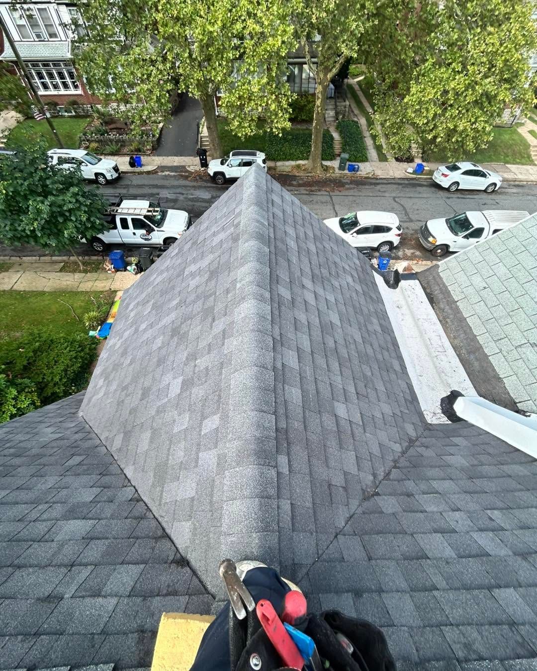 Worker on a shingled roof, looking down over a street with parked cars and trees.