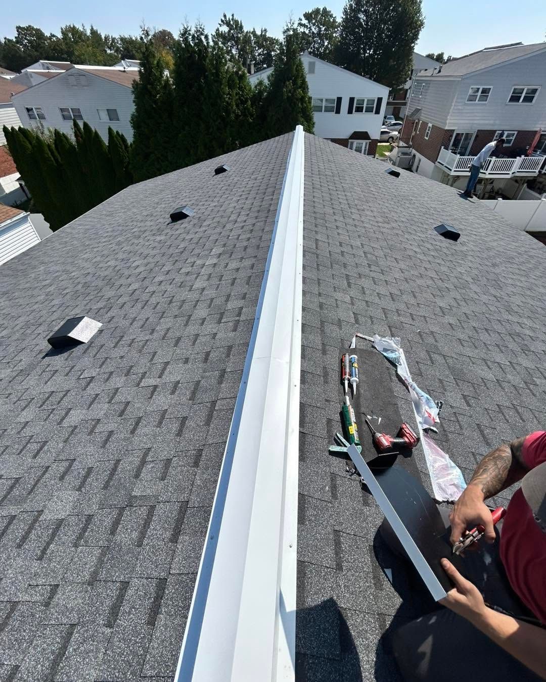 Roofer working on a gray shingle roof along a central ridge vent, with houses and trees in the background