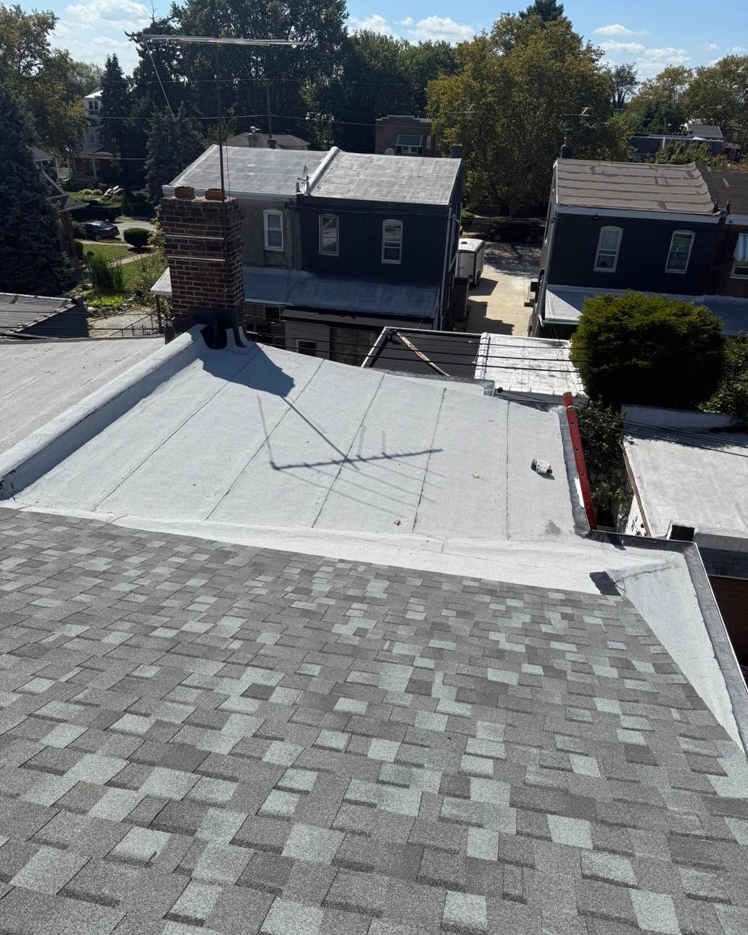 Rooftop with gray shingles and white flat roof, overlooking two dark houses and trees in a neighborhood.