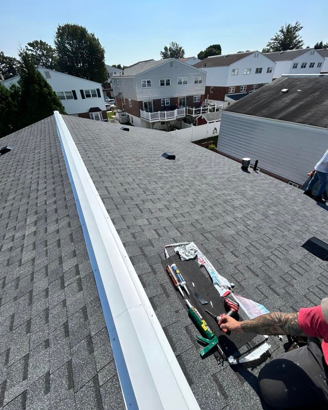 Worker on a gray shingled roof beside white siding, using tools near a roof seam.