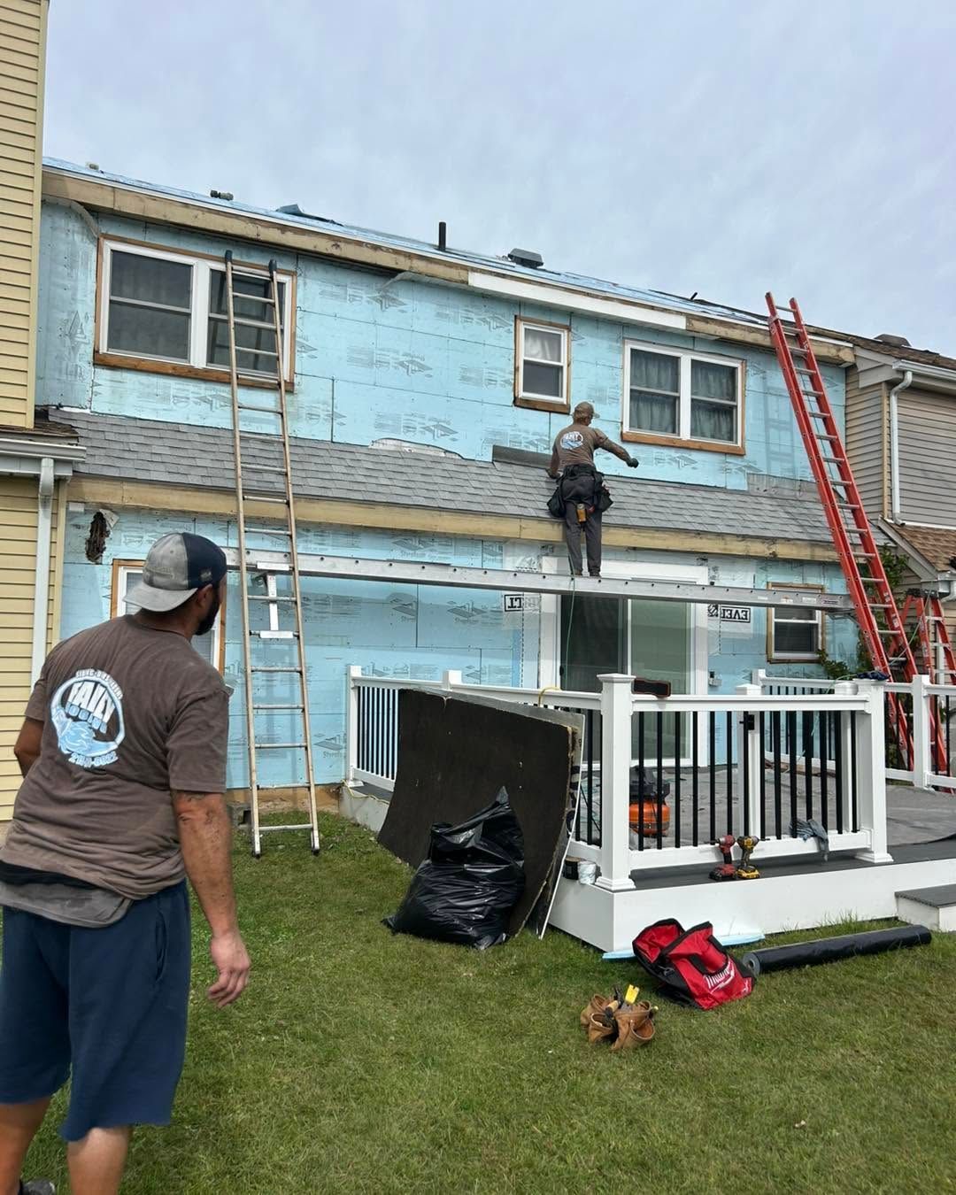 Workers painting the upper exterior of a blue townhouse, with ladders and tools in the yard