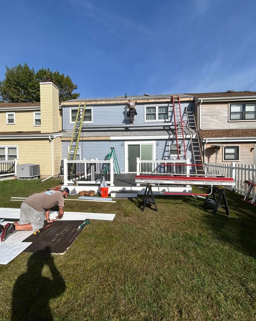 Workers reroofing a townhouse row, with ladders, scaffolding, and roofing materials on the lawn.