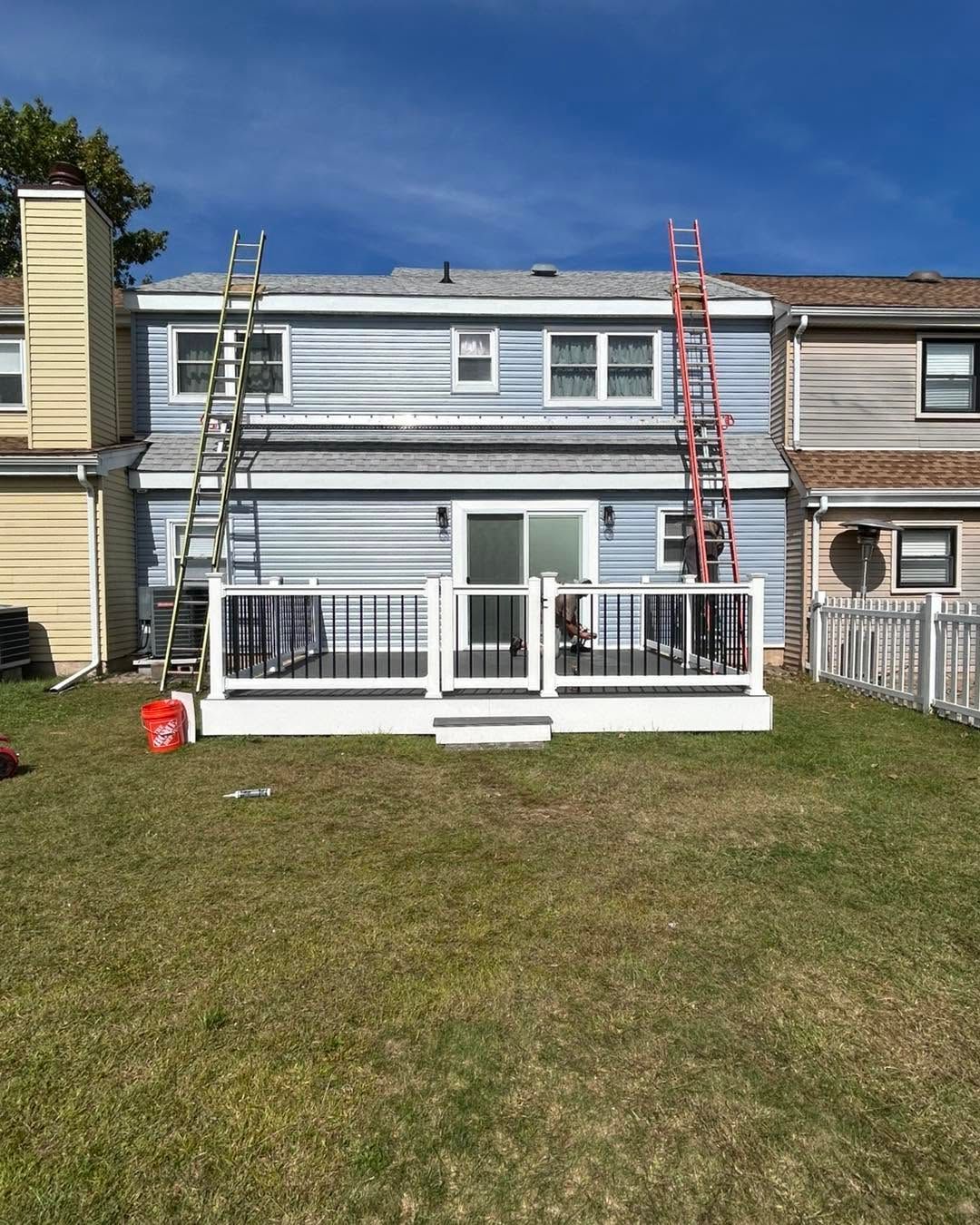 Two-story house with white balcony railings and ladders in front, viewed from a grassy yard.