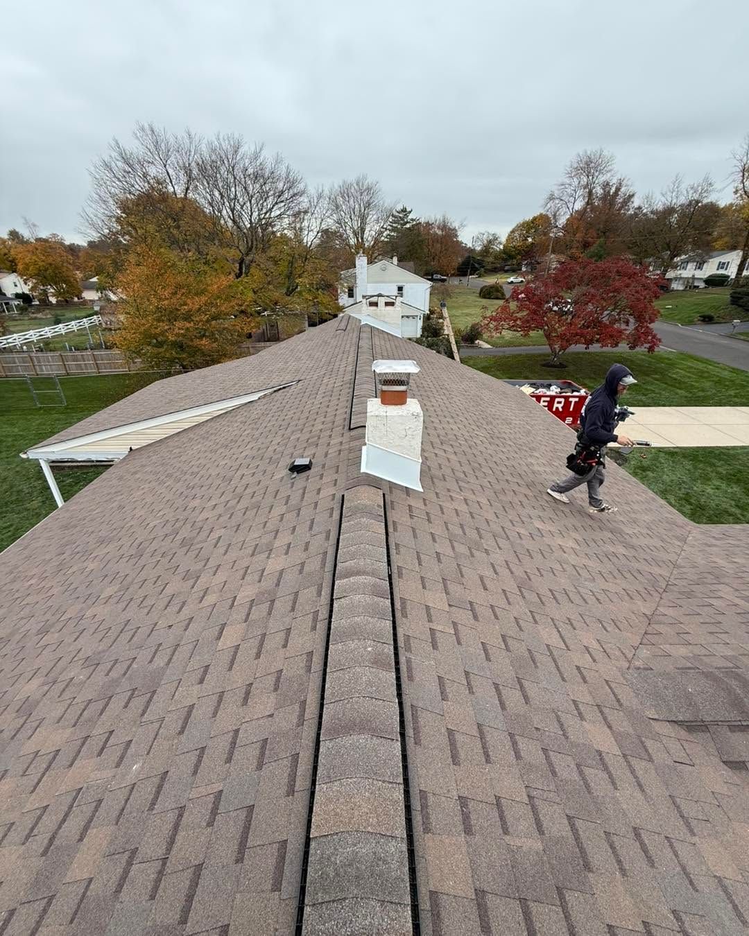 Rooftop view with shingles, chimney vents, and a worker on the right near autumn trees.