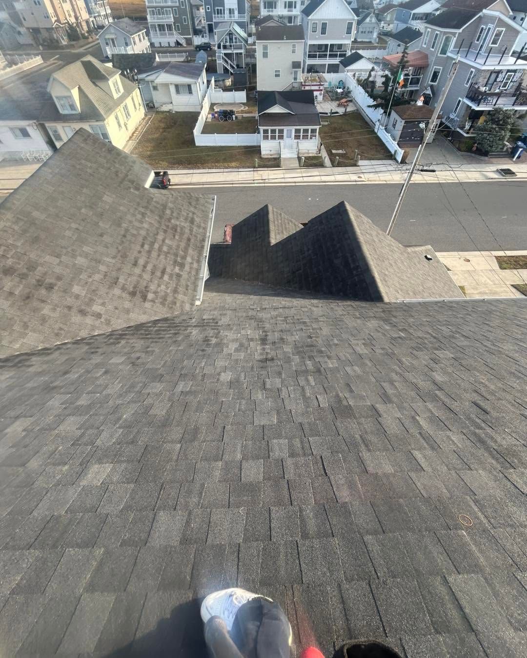 View from a rooftop over gray shingles toward a suburban street with houses and a driveway below