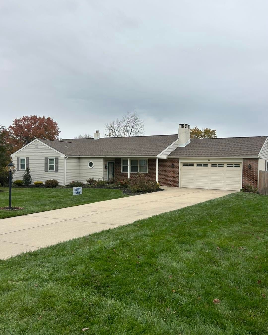 Single-story suburban house with attached garage, long driveway, and green lawn under cloudy sky