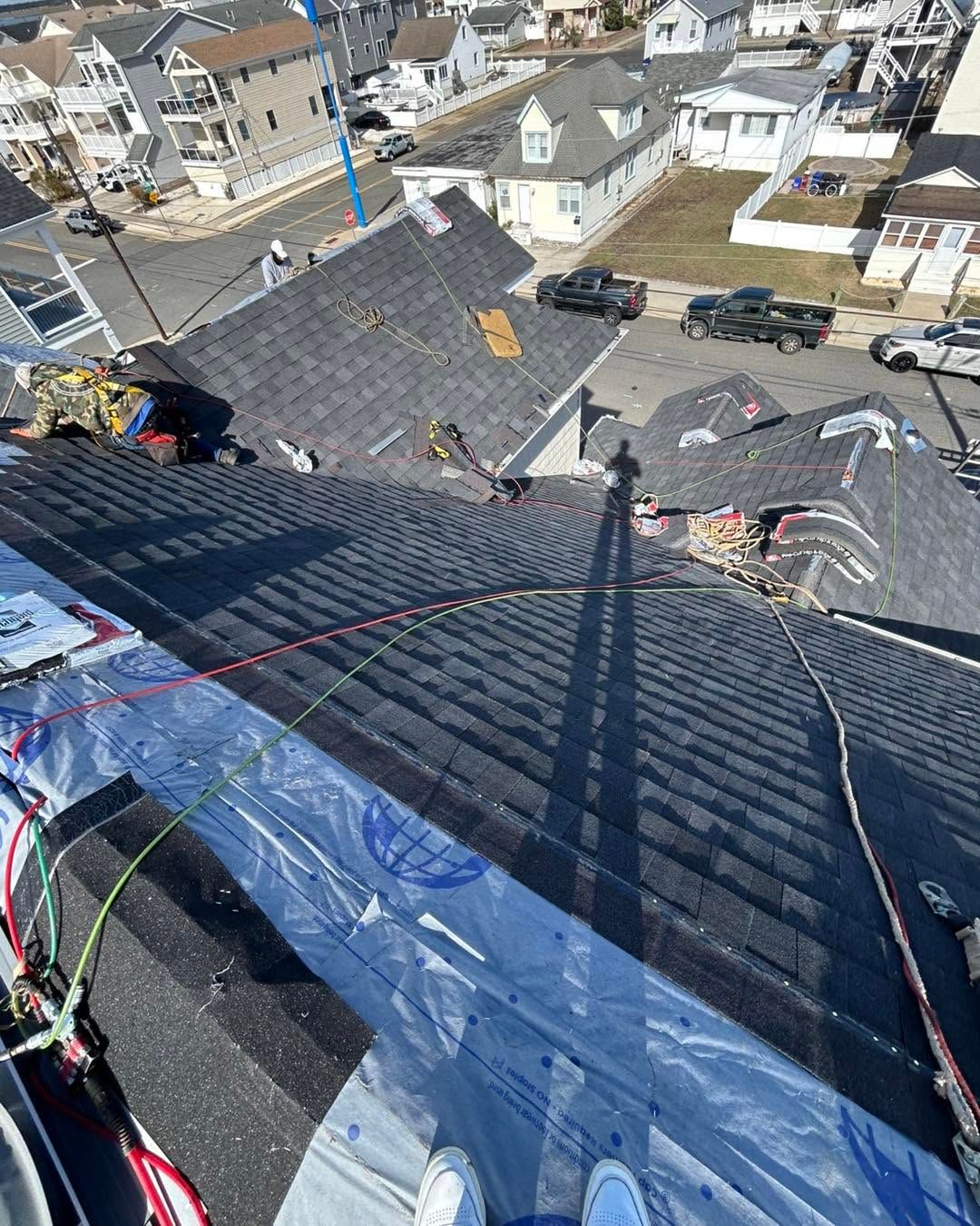 Rooftop under repair with dark shingles, blue tarp, ropes, and houses visible below.