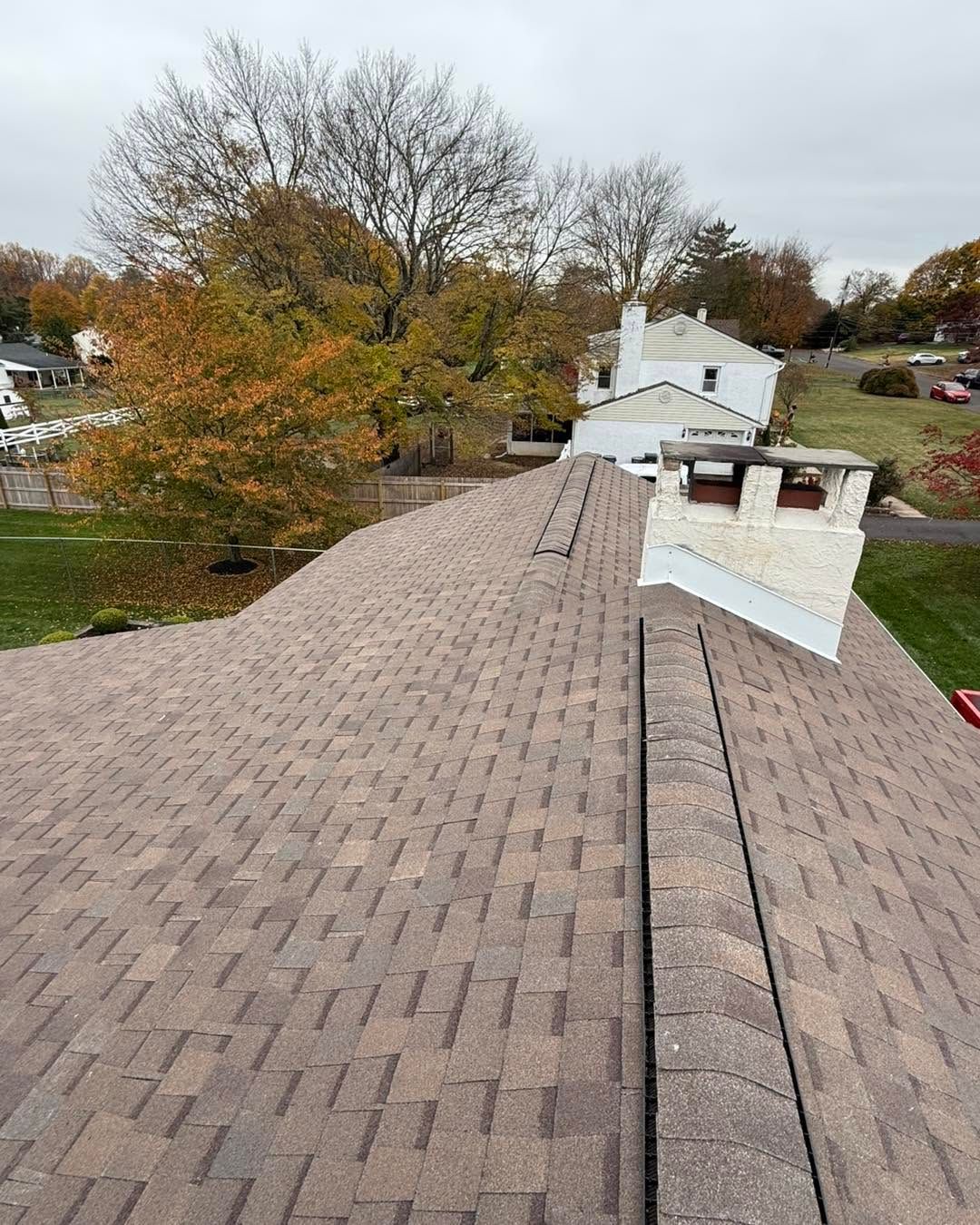 Rooftop view of a shingled house with a chimney, trees, and neighboring homes in a suburban area.