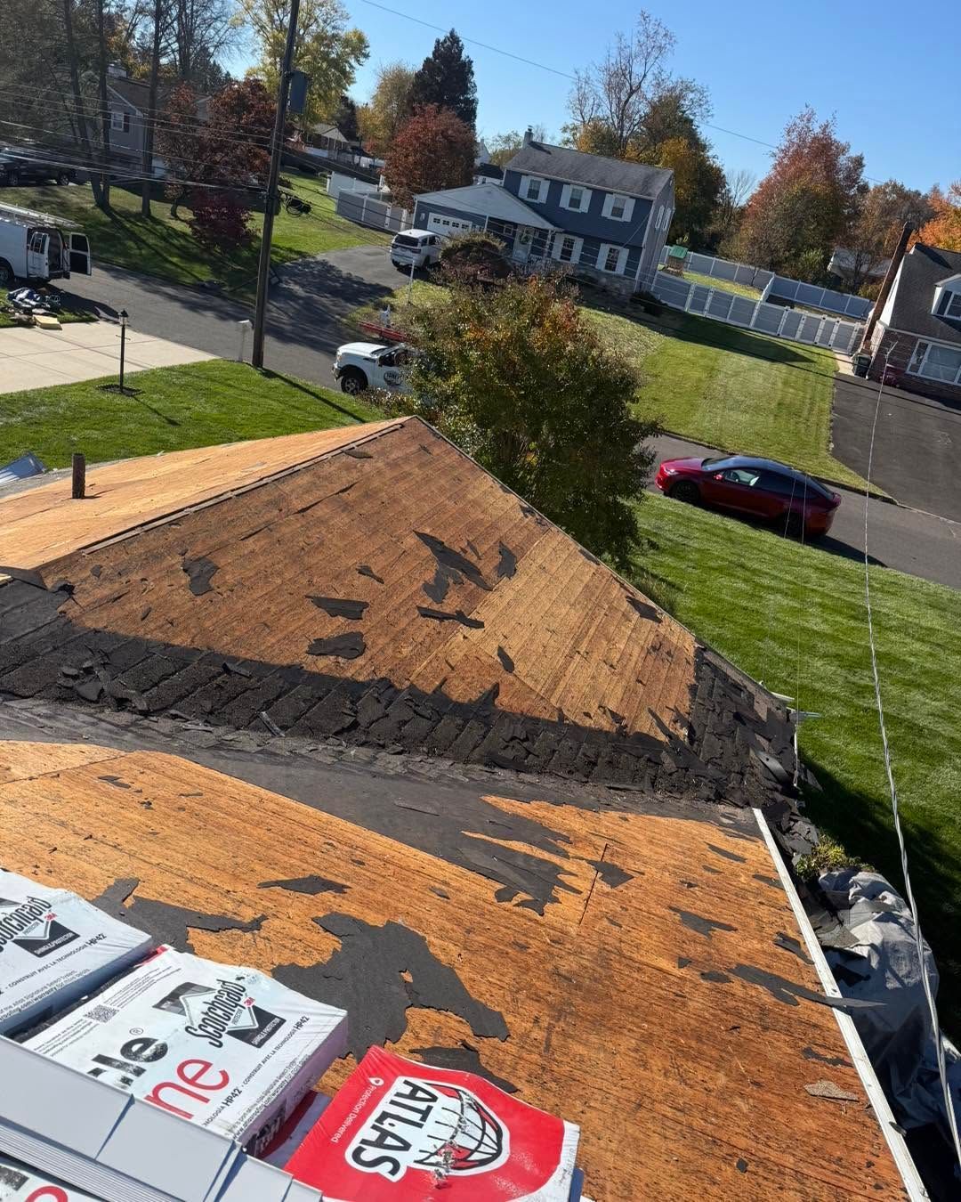 Rooftop with exposed shingles, roofing materials, and a suburban street with houses and parked cars below