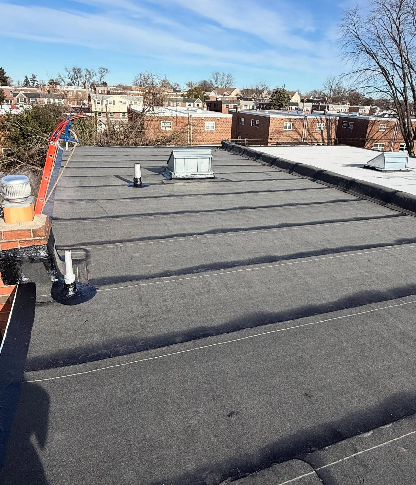 Flat rooftop with black roofing, vents, and nearby houses under a clear blue sky.