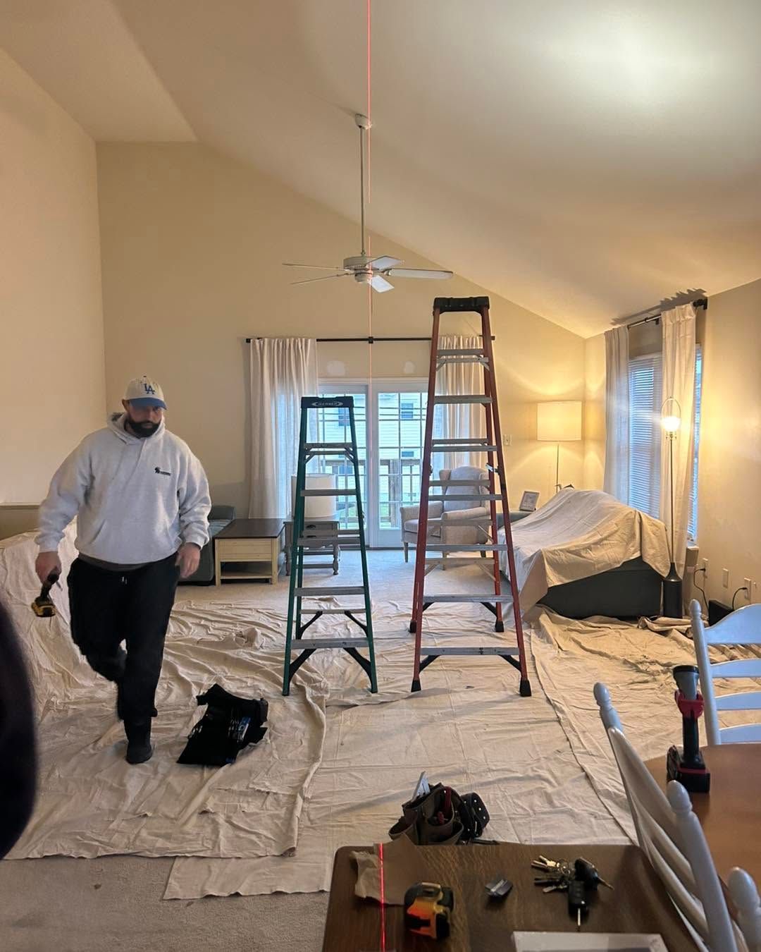 Person in a room covered with drop cloths, with ladders, tools, and a painted ceiling in progress.