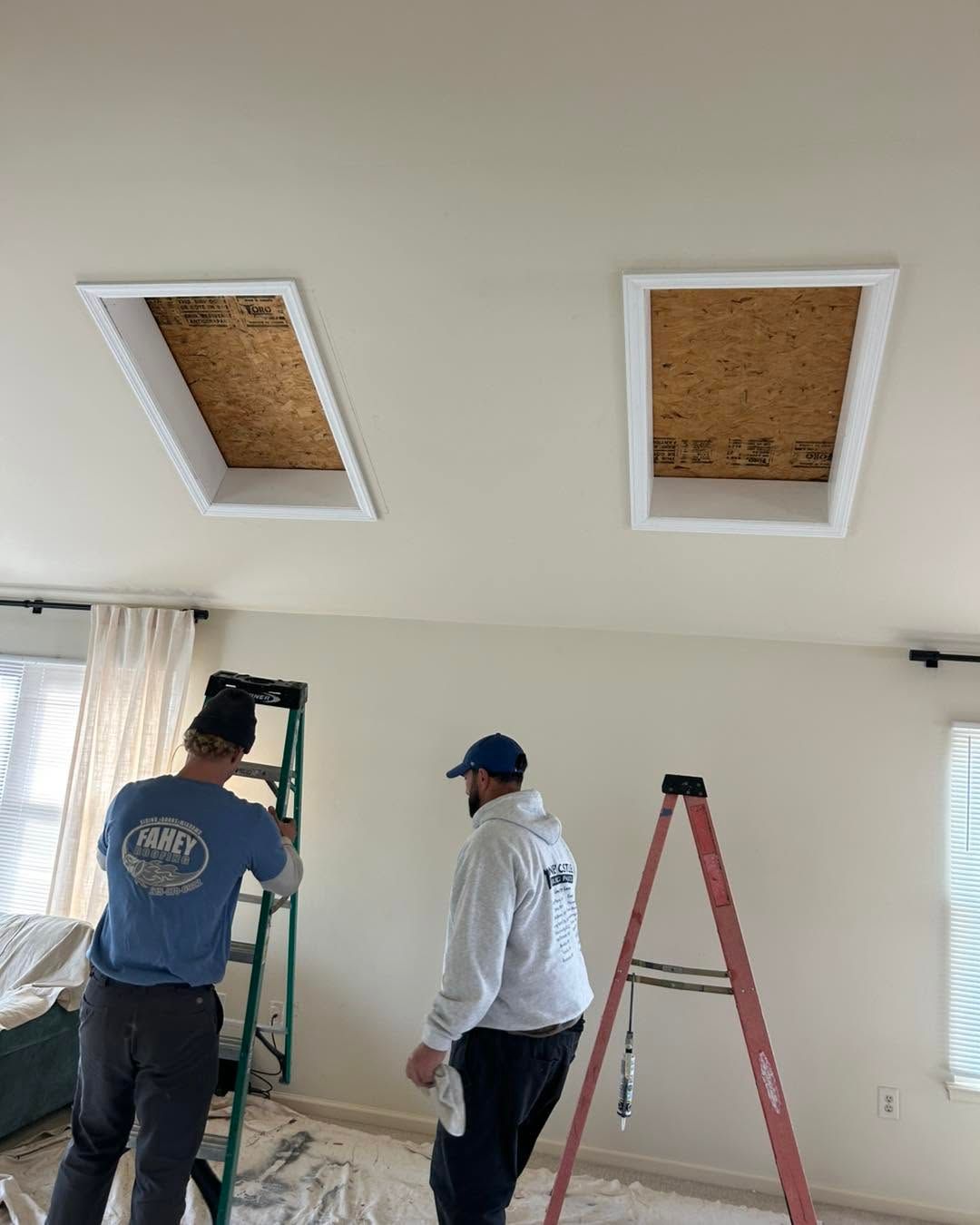Two workers on ladders inspect two open ceiling panels in a bright room.