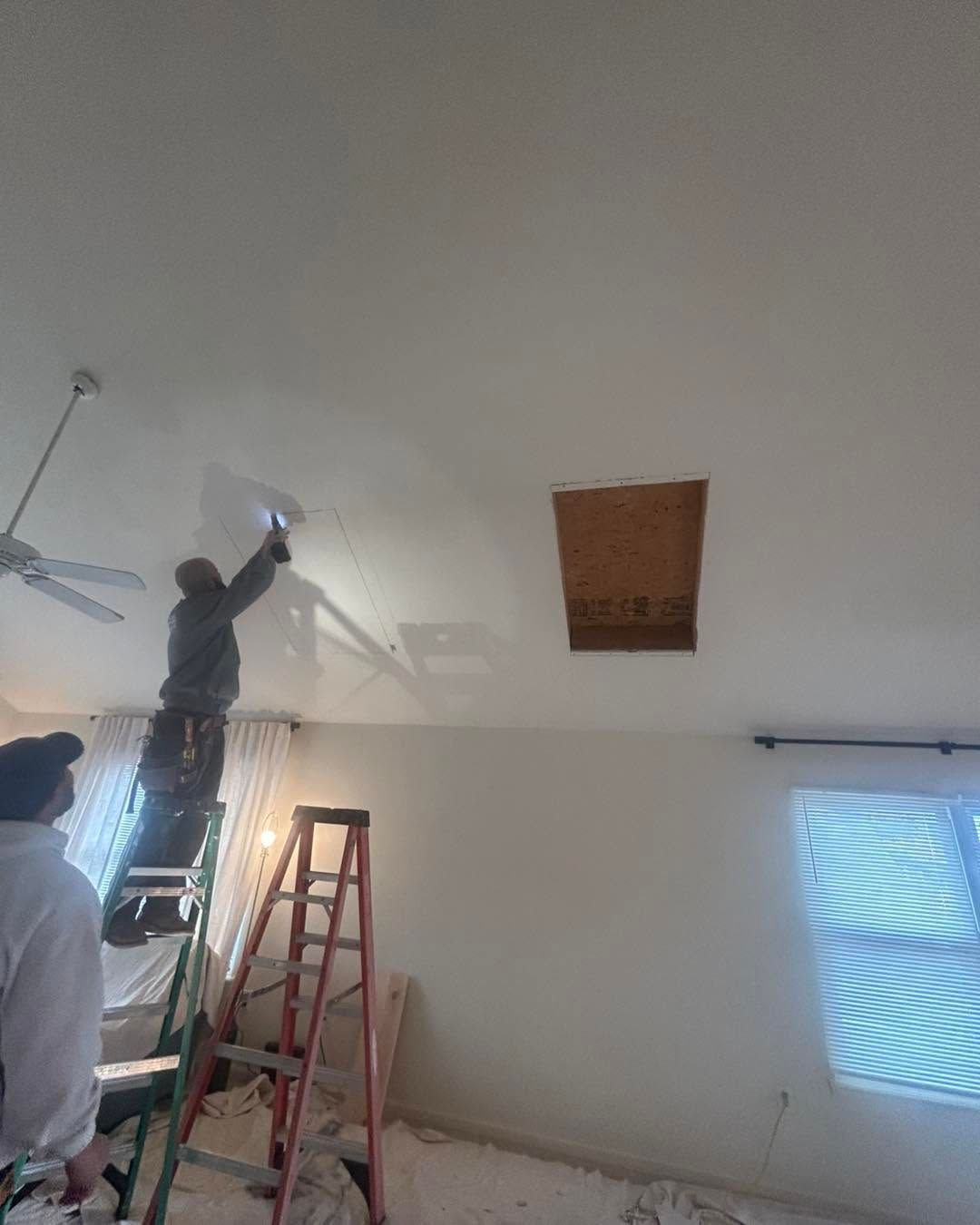 Workers painting a white ceiling indoors, with a ladder and a window on the right.
