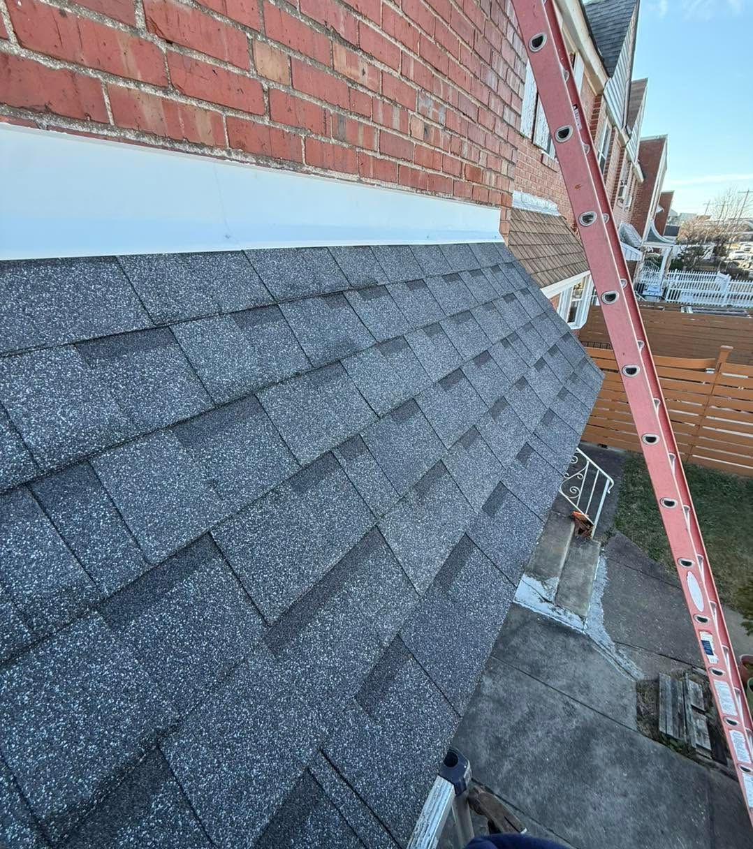Roof shingles under repair beside a brick wall, with a red ladder leaning against the building.