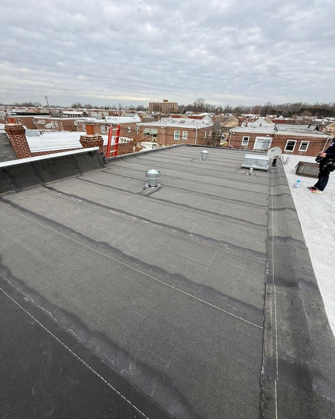 Flat rooftop with dark gray roofing and snow, overlooking a snowy city skyline under cloudy skies.