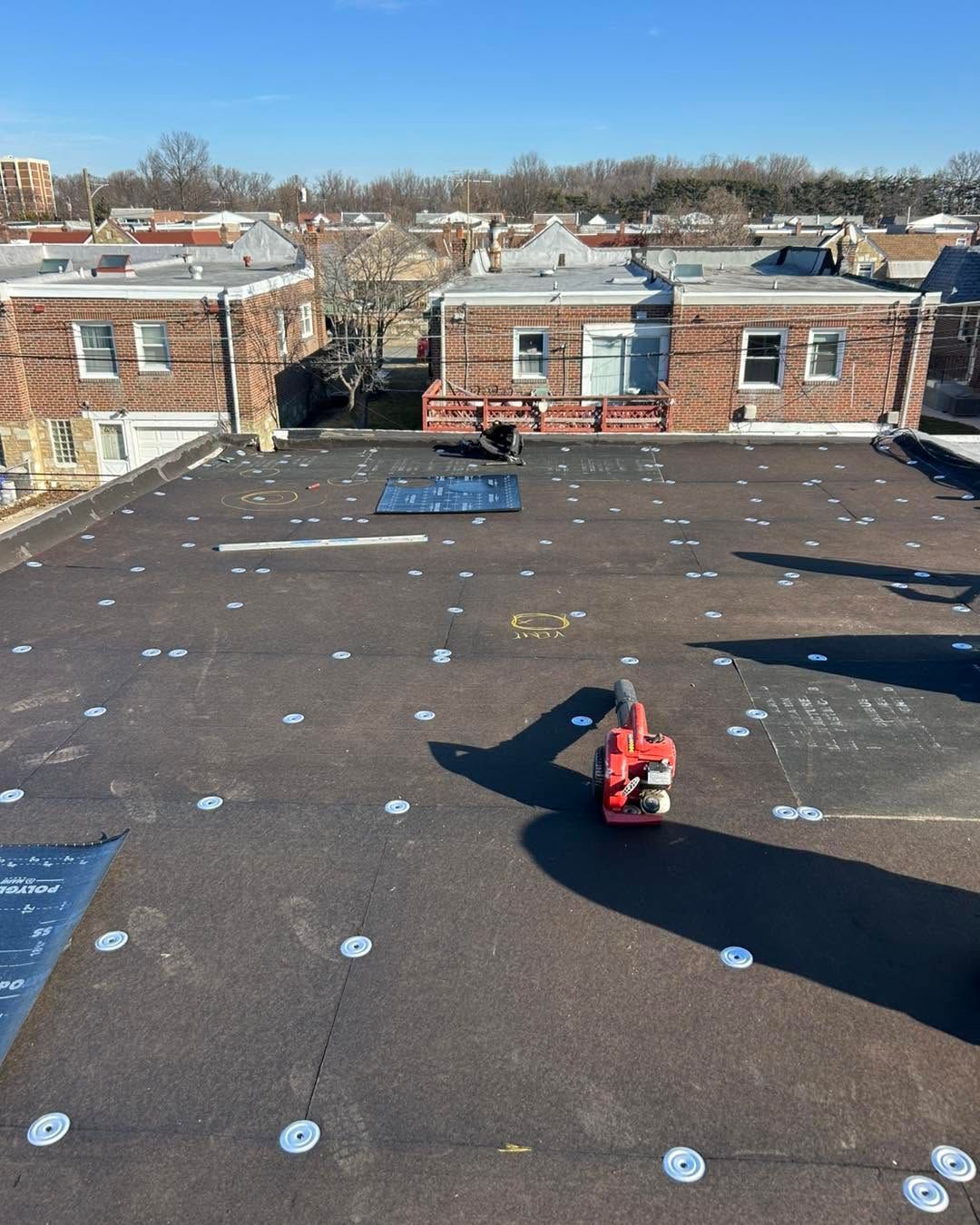 Rooftop with black roofing membrane, white fasteners, and a red tool near the center under a clear blue sky.