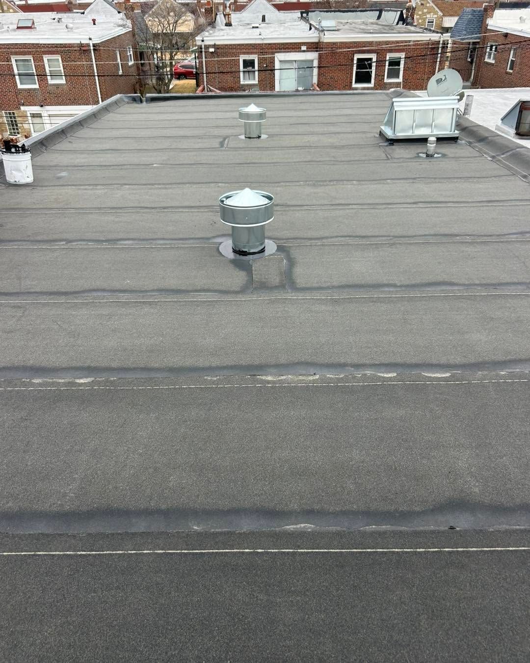 Flat rooftop with gray tar paper, two vents, and nearby brick buildings in the background.
