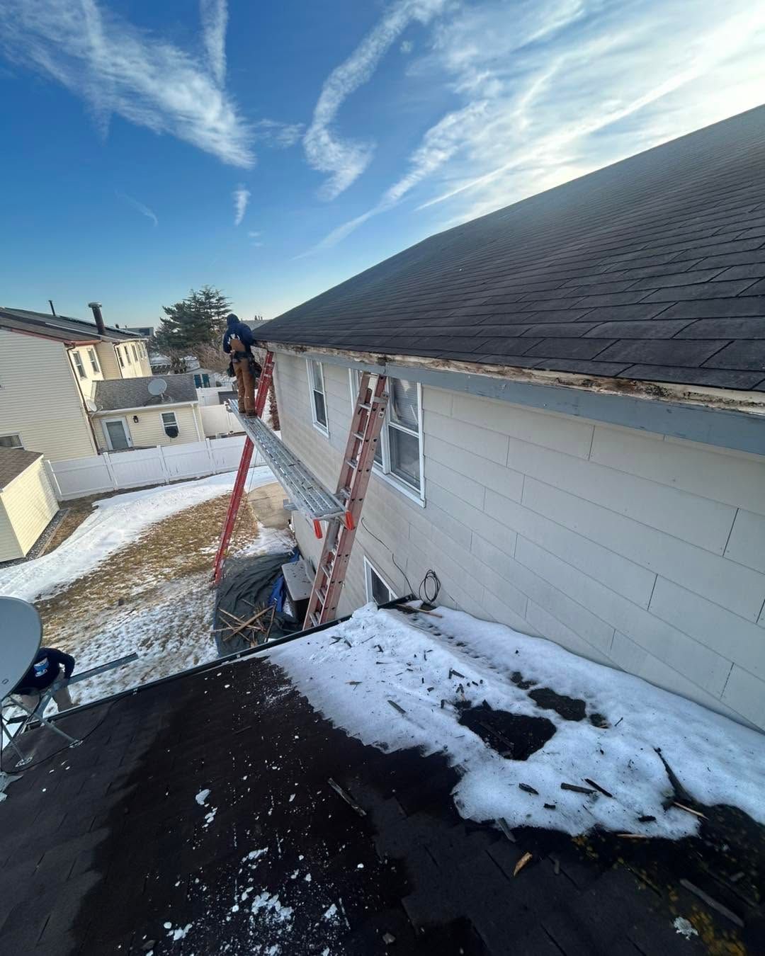 Snow-covered roof with two ladders leaning against a house under a blue sky.