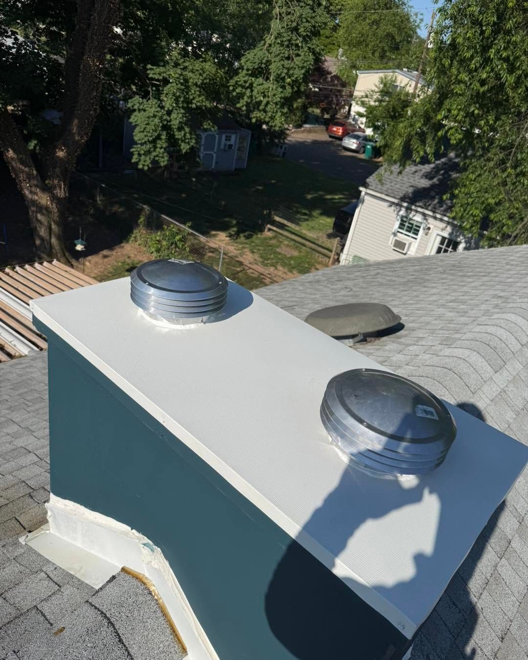 Rooftop vent boxes with silver caps on a shingled roof, with trees and houses in the background