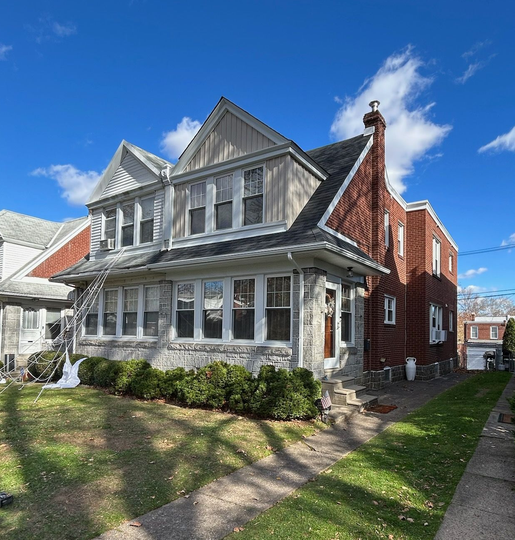 Sunny street view of a gray Victorian house with white trim, red brick side, and a front porch under blue sky