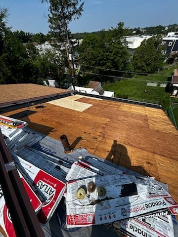 Roof under construction with plywood sheathing, tools, and roofing paper in a sunny suburban neighborhood.