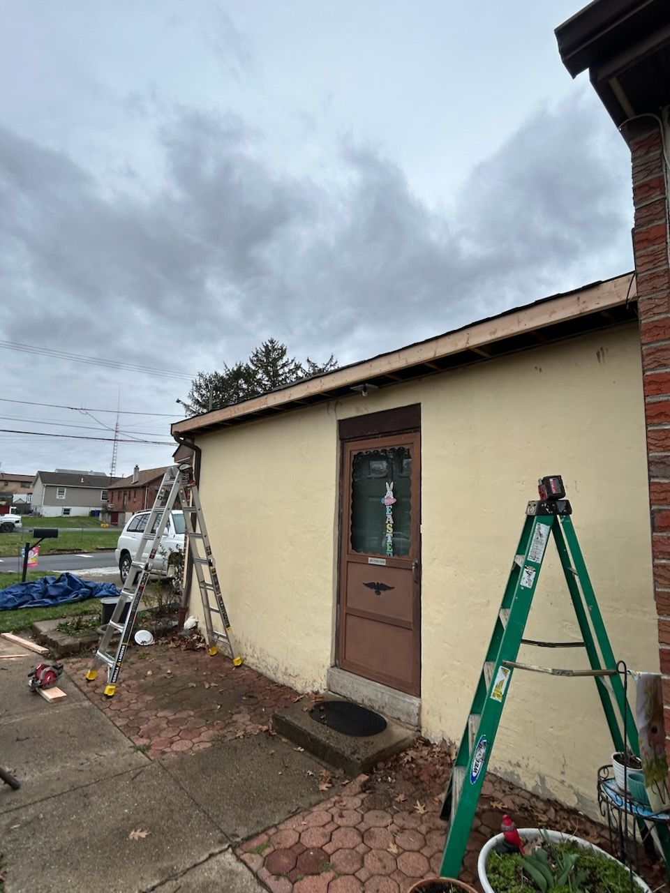 Small beige house exterior with a brown door, ladders, and a green step ladder under a cloudy sky.