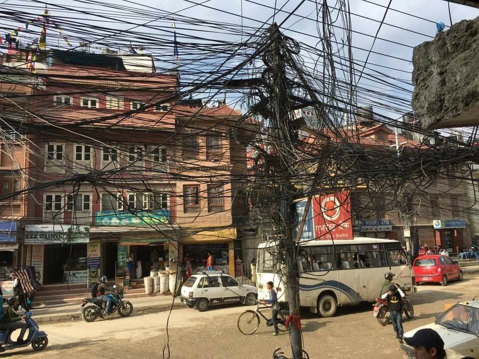 Busy city street with brick buildings, tangled overhead wires, pedestrians, motorcycles, and parked cars
