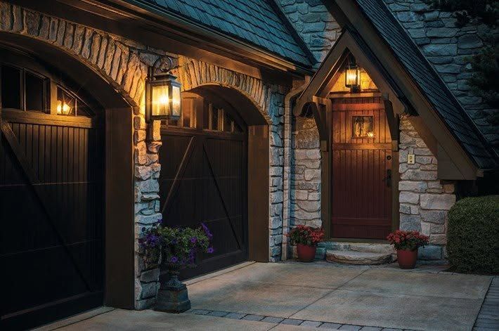 Warmly lit stone cottage entrance with wooden doors, arched porch, and potted flowers at dusk