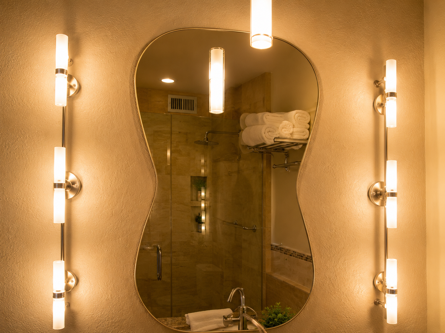Backlit bathroom mirror with wall sconces and a shower reflected in warm light