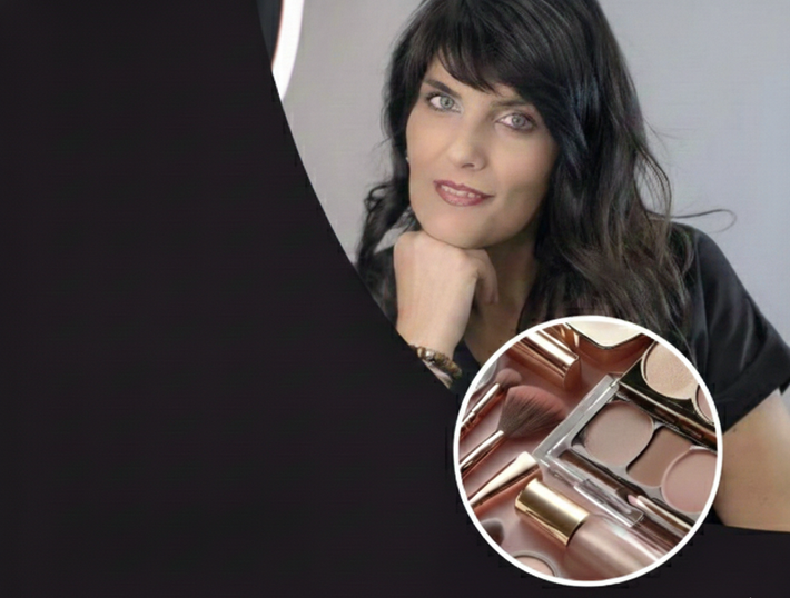 Donna con capelli scuri, mento appoggiato sulla mano, che guarda verso la telecamera; prodotti per il trucco in primo piano.