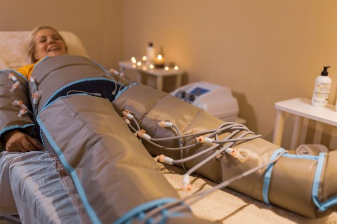 Donna sdraiata su un letto con maniche a compressione, sorridente. Ambiente spa con candele e attrezzature.