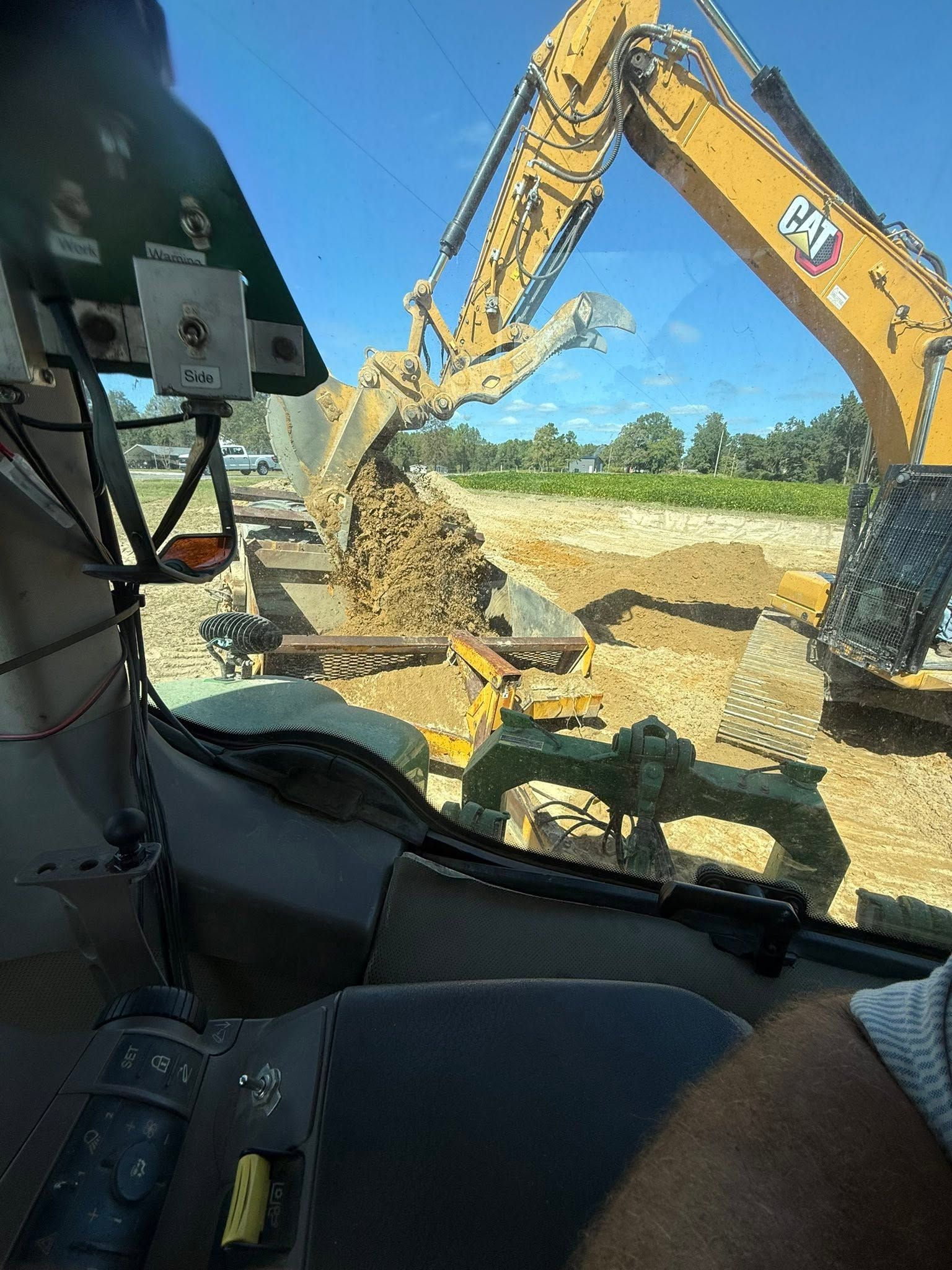 Excavator arm digging dirt from inside a construction vehicle cab, with blue sky and field outside.
