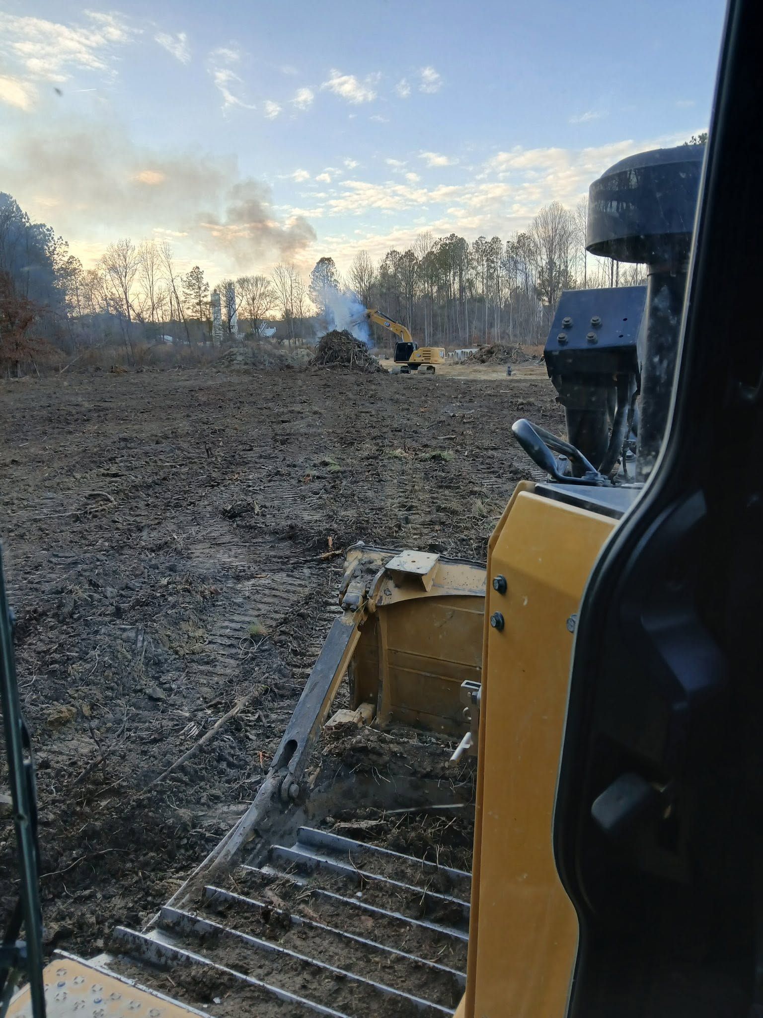 Yellow excavator working in a muddy field at sunrise, with trees and smoke in the distance.