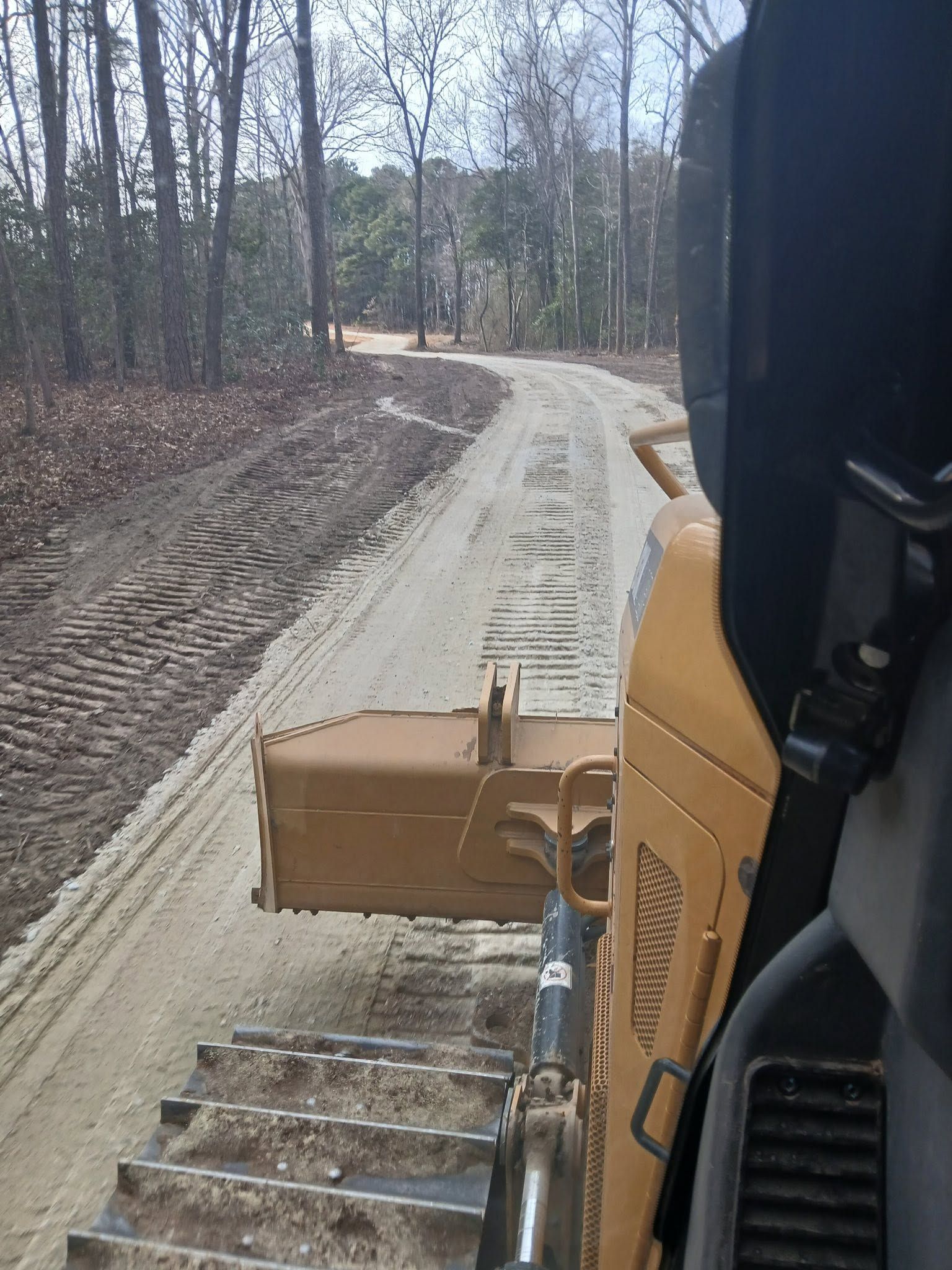 Excavator blade pushing a cardboard box along a sandy dirt road in a wooded construction area