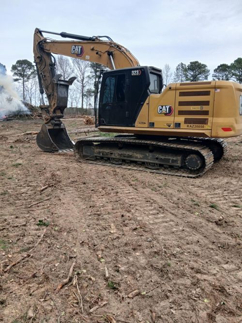 Yellow excavator on a muddy construction site, bucket raised over rough dirt and trees.