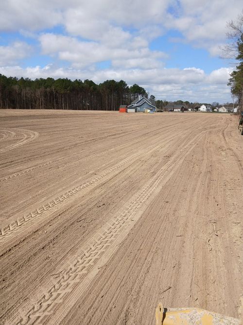 Freshly cleared dirt lot under a cloudy sky, with trees and distant houses along the horizon.