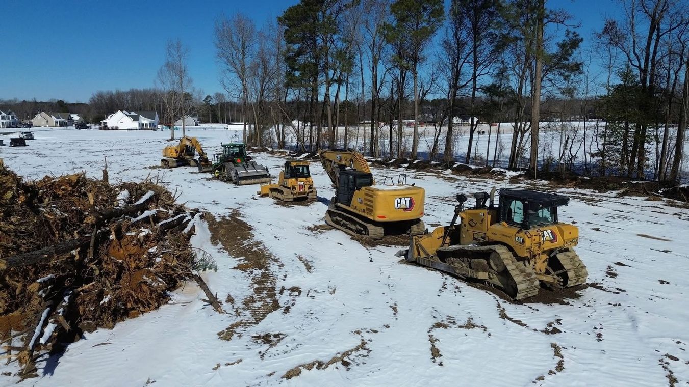 Yellow bulldozers lined up on a snowy construction site beside a muddy ditch and bare trees