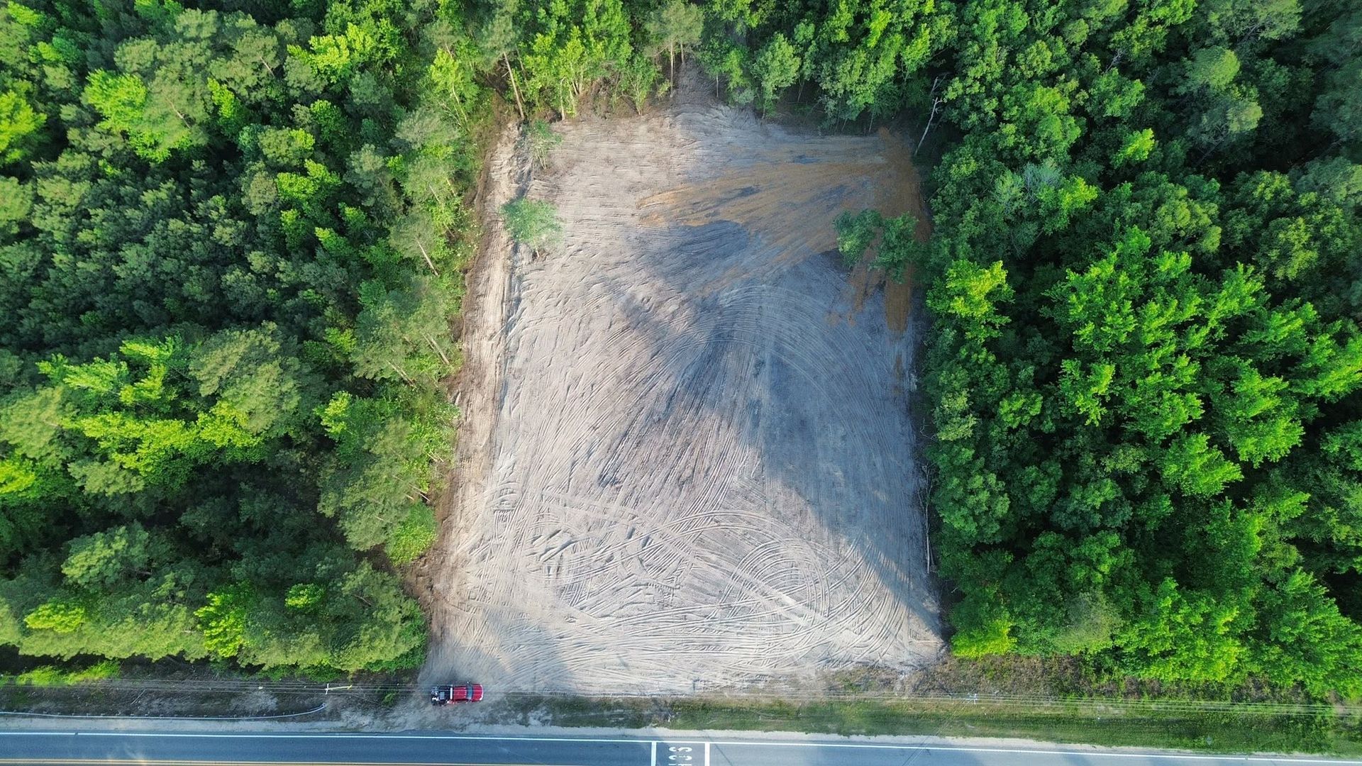Aerial view of a gravel clearing surrounded by dense green trees beside a road.