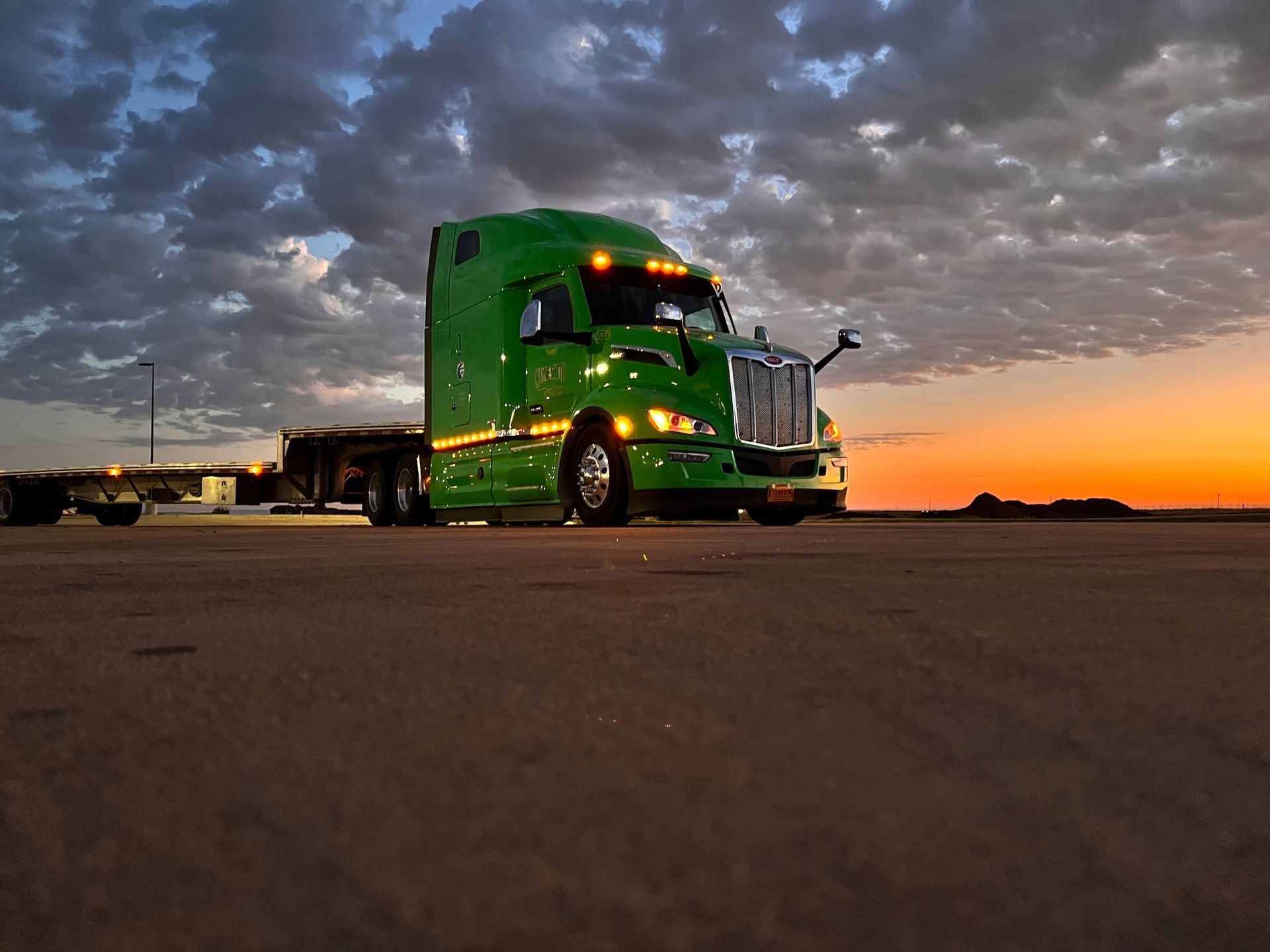 Green semi-truck parked on a road at sunset under dramatic cloudy skies