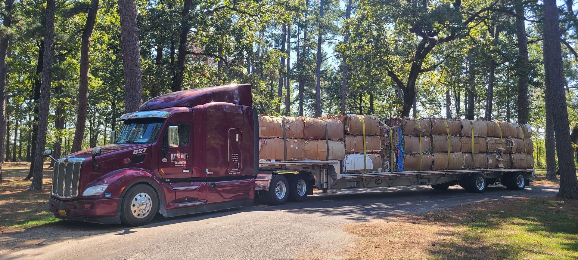 Maroon semi-truck hauling a long trailer loaded with stacked logs in a wooded area