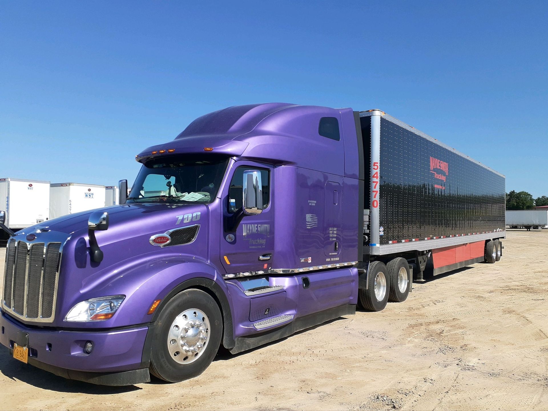 Purple semi truck with black trailer parked on a dirt lot under a blue sky