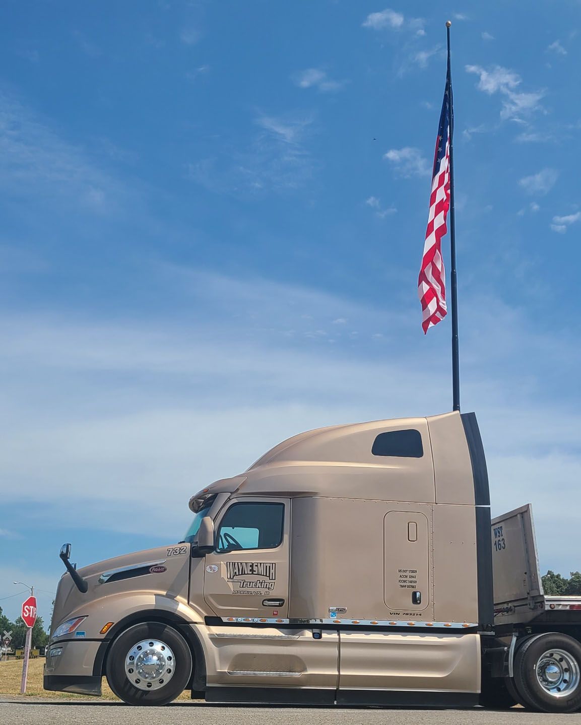 Tan semi-truck parked under a tall flagpole with a waving American flag against a blue sky