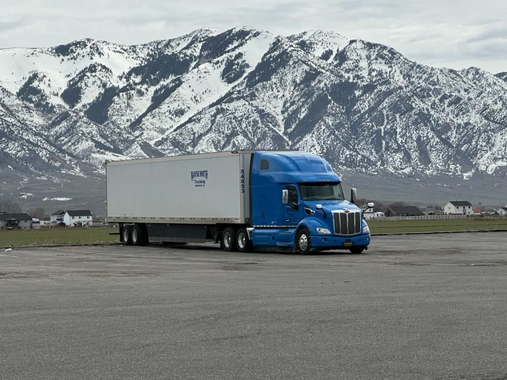 Blue semi-truck with white trailer parked on a lot, snowy mountains in the background.