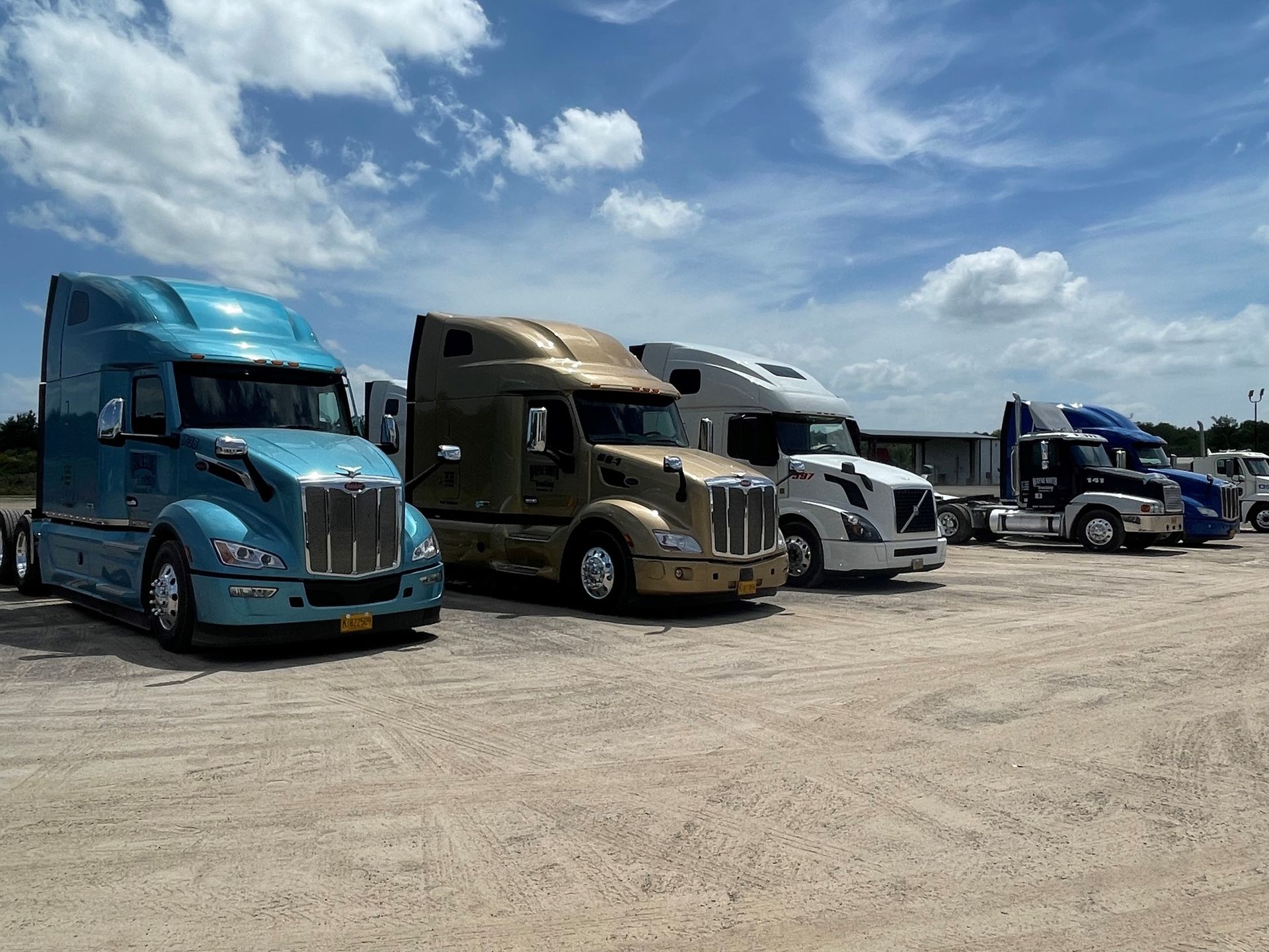 Several parked semi-trucks in a gravel lot under a partly cloudy sky