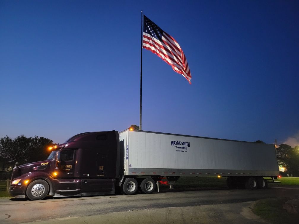 Semi-truck with trailer parked at dusk beneath a large American flag on a tall pole.