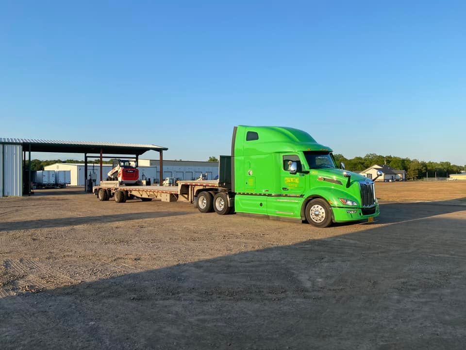 Bright green semi-truck parked on a gravel lot beside an open-sided shed with a flatbed trailer.