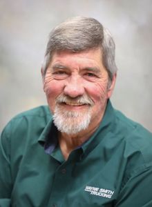 Smiling man in a green shirt with gray hair and beard against a soft gray background