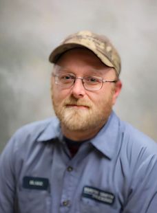 Man in a camouflage cap and blue uniform shirt, wearing glasses, against a gray backdrop.