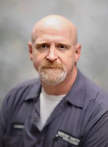 Portrait of a bald man with a goatee in a gray shirt, facing the camera against a soft gray background