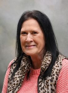Smiling woman with long dark hair wearing a pink top and leopard-print scarf against a gray background